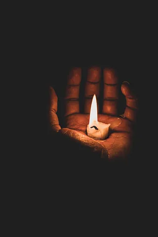 In der Dunkelheit ist eine Hand zu sehen. Die nach oben geöffnete Hand hält eine heruntergebrannte Kerze, die die Hand beleuchtet.