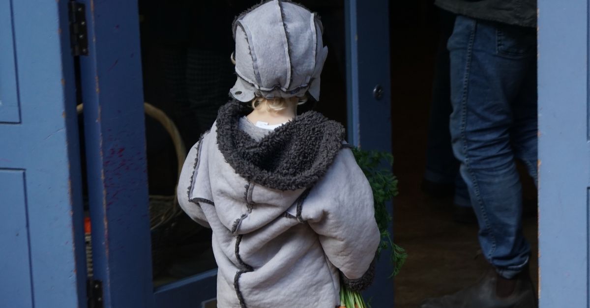 Ein kleines Kind mit Mütze und Jacke geht über eine Türschwelle in ein Haus. In seiner Hand hält es einen Bund Möhren.