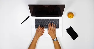 Hände tippen auf einer Laptop-Tastatur, daneben liegen Stift, Orange und Smartphone auf weißem Tisch.