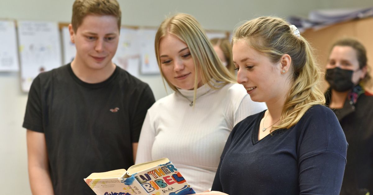 Drei Jugendliche stehen beieinander und schauen gemeinsam in eine Bibel, die eine der Jugendlichen in der Hand hält.