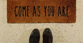 Zwei Füße in dunklen Schuhen vor einer Fußmatte an einer Tür. Auf der Fußmatte steht geschrieben "Come as you are".
