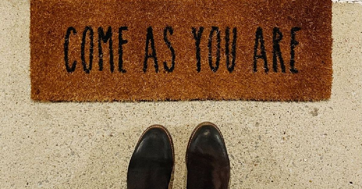 Zwei Füße in dunklen Schuhen vor einer Fußmatte an einer Tür. Auf der Fußmatte steht geschrieben "Come as you are".