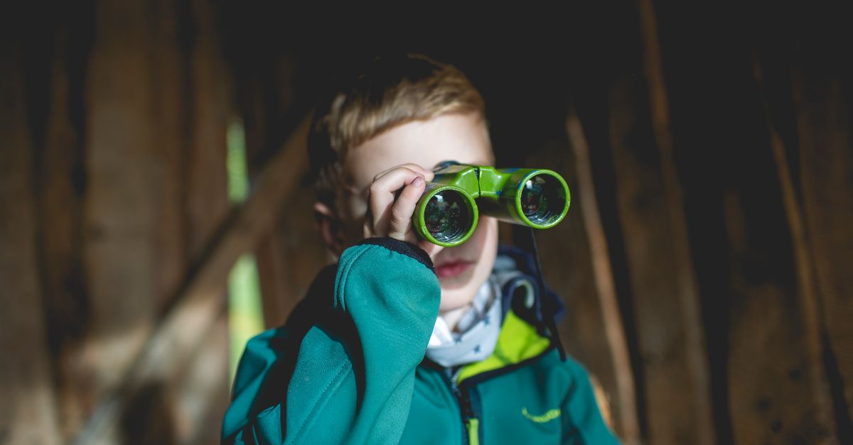 Ein Junge vor einer Holzwand hält ein Fernglas vor die Augen.