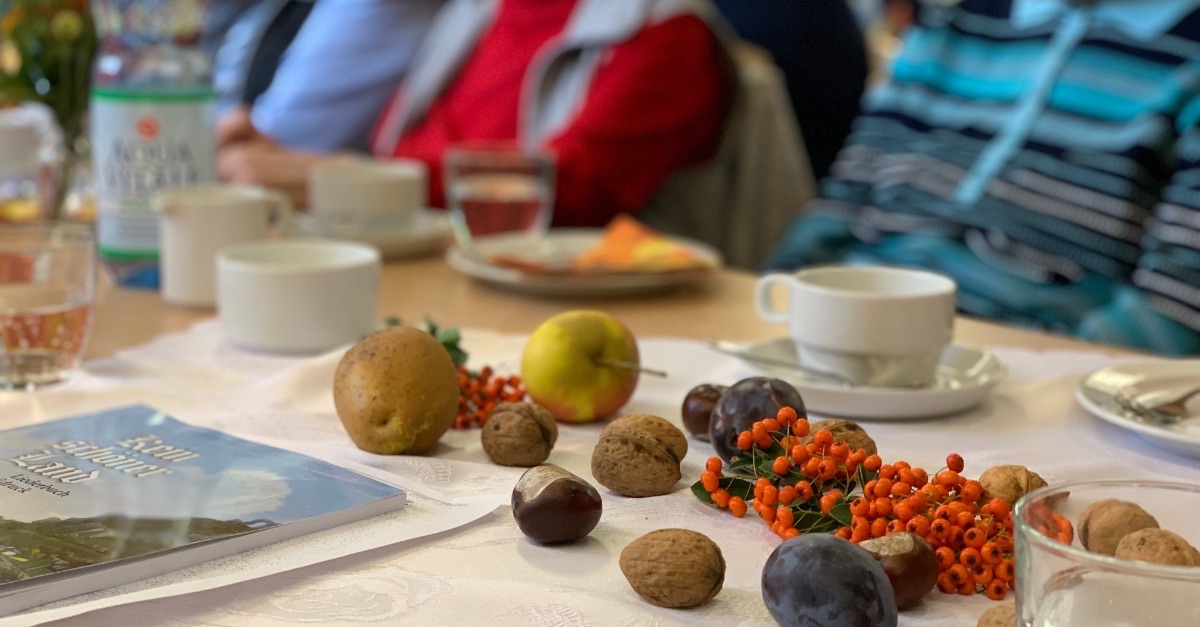 Ein Tisch mit Herbstdekoration aus Nüssen, Pflaumen und roten Beeren. Auf dem Tisch stehen Kaffeetassen. Im Hintergrund unscharfe ältere Menschen bei einer Zusammenkunft.
