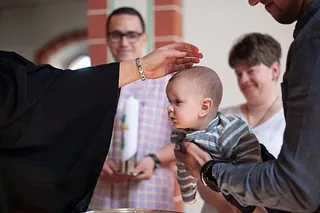 Ein Baby wird bei einer Taufe von einer Person im Talar gesegnet, während zwei Erwachsene im Hintergrund zuschauen.