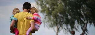 Ein Mann trägt zwei kleine Kinder in einem Mohnblumenfeld, ein weiteres Kind läuft voraus. Ein Baum steht im Hintergrund.