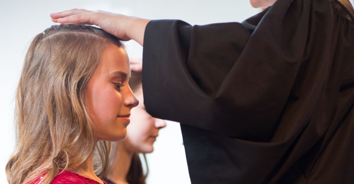 Eine Konfirmandin in festlichem, rotem Kleid kniet, während eine Pfarrerin ihr die Hände auflegt und sie segnet. Neben ihr im Bildhintergrund kniet eine weitere Konfirmandin.