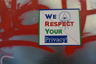 An einer Wand mit Graffiti ist ein Aufkleber befestigt. Er zeigt ein stilisiertes Auge, darunter der Schriftzug "We respect your privacy".