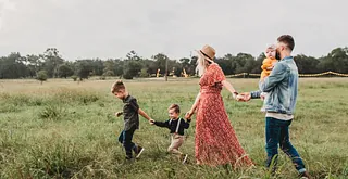 Eine Familie geht über ein Feld im Sommer. Die Eltern und die drei Kinder halten sich dabei an den Händen.