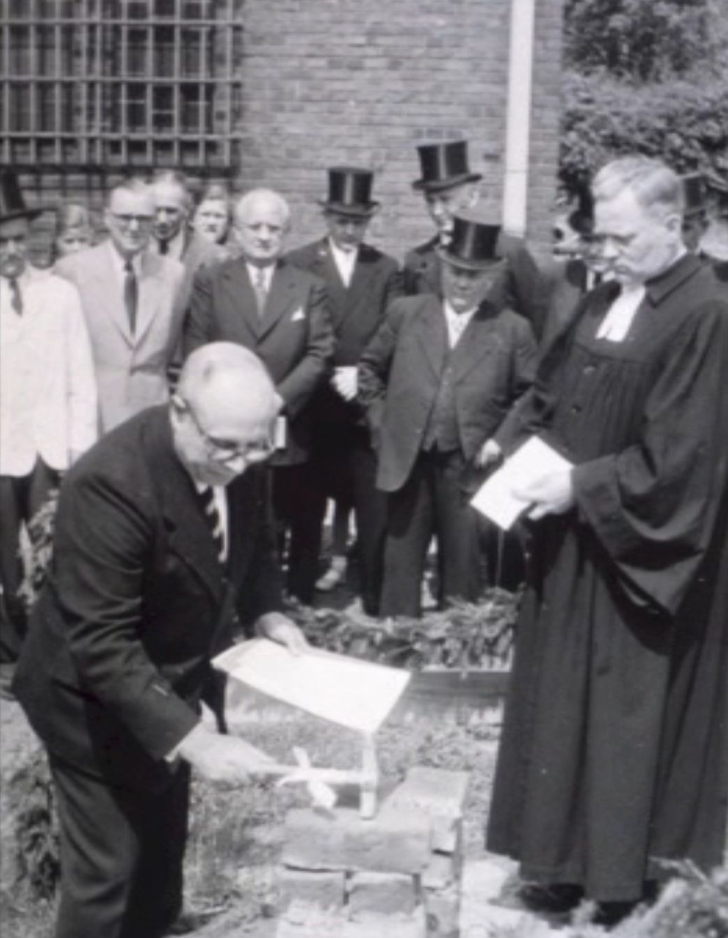 Ein Mann haut mit einem Hammer auf den Grundstein des Kindergartens vor Baubeginn. Er ist umgeben von Männern in Anzügen und Zylindern, neben ihm steht ein Pfarrer, dieser hält ein Buch in den Händen.