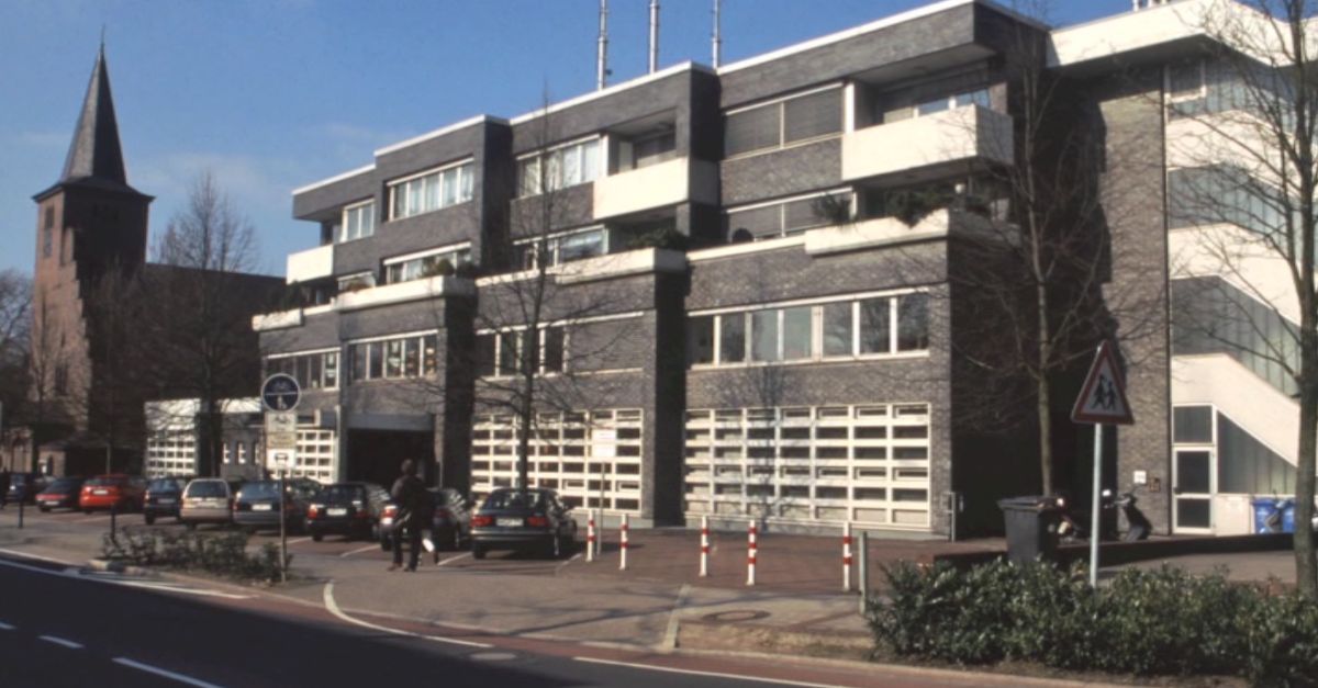 Mehrstöckiges, modernes Gemeindehaus mit großen Fenstern und mehreren geparkten Autos davor, im Hintergrund der Kirchturm.