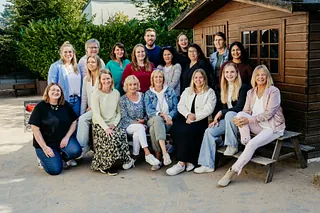 Gruppenfoto des Kindergarten-Teams 2024, 17 Personen, die draußen vor einem Holzhaus sitzen und stehen, lächelnd und in lässiger Kleidung.