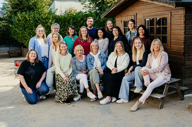 Gruppenfoto des Kindergarten-Teams 2024, 17 Personen, die draußen vor einem Holzhaus sitzen und stehen, lächelnd und in lässiger Kleidung.