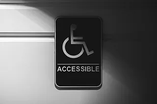 Auf grauer Wand hängt ein Schild. Es zeigt einen stilisierten Rollstuhlfahrer, darunter der Schriftzug "Accessible".