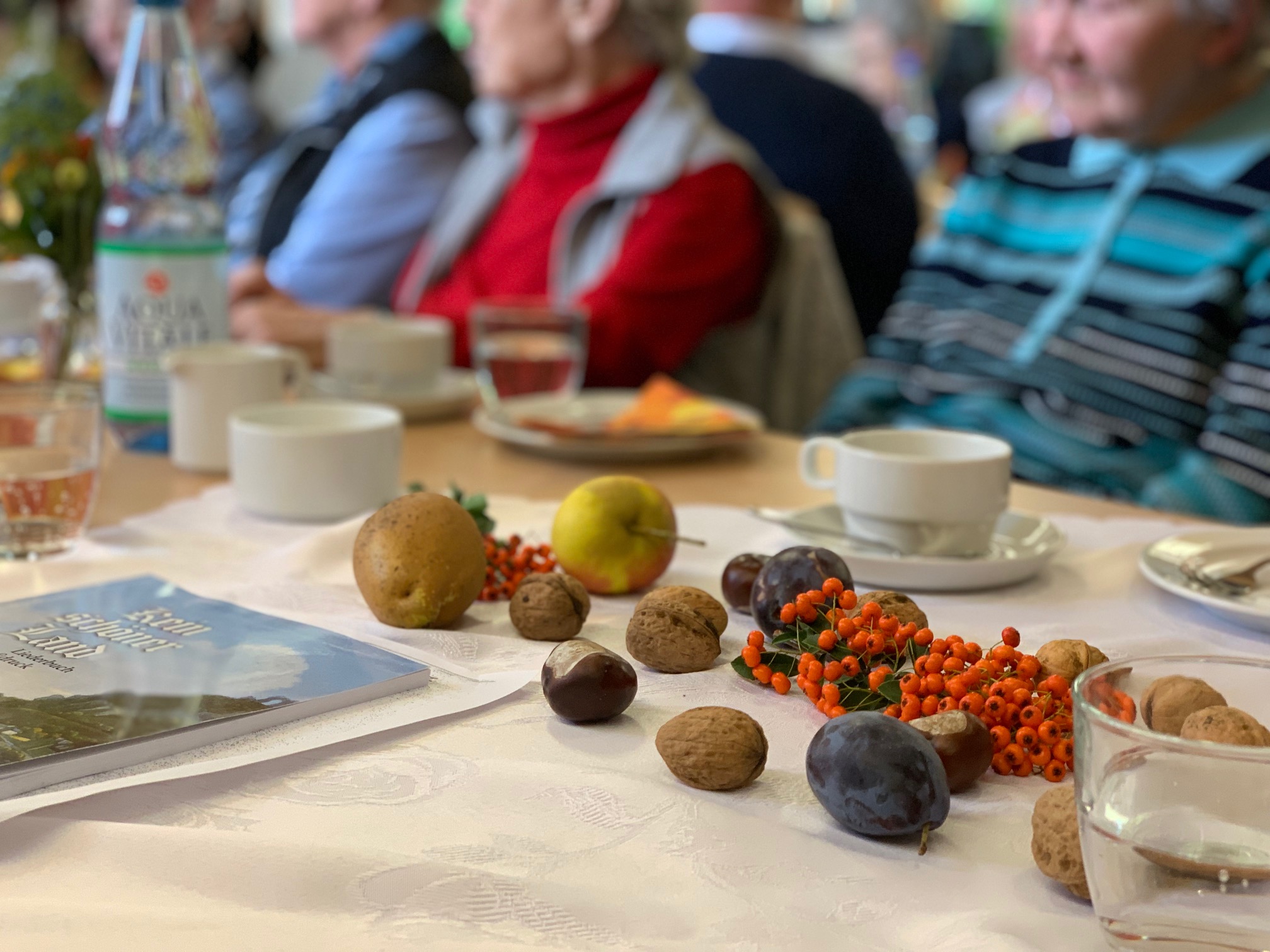 Ein Tisch mit Herbstdekoration aus Nüssen, Pflaumen und roten Beeren. Auf dem Tisch stehen Kaffeetassen. Im Hintergrund unscharfe ältere Menschen bei einer Zusammenkunft.