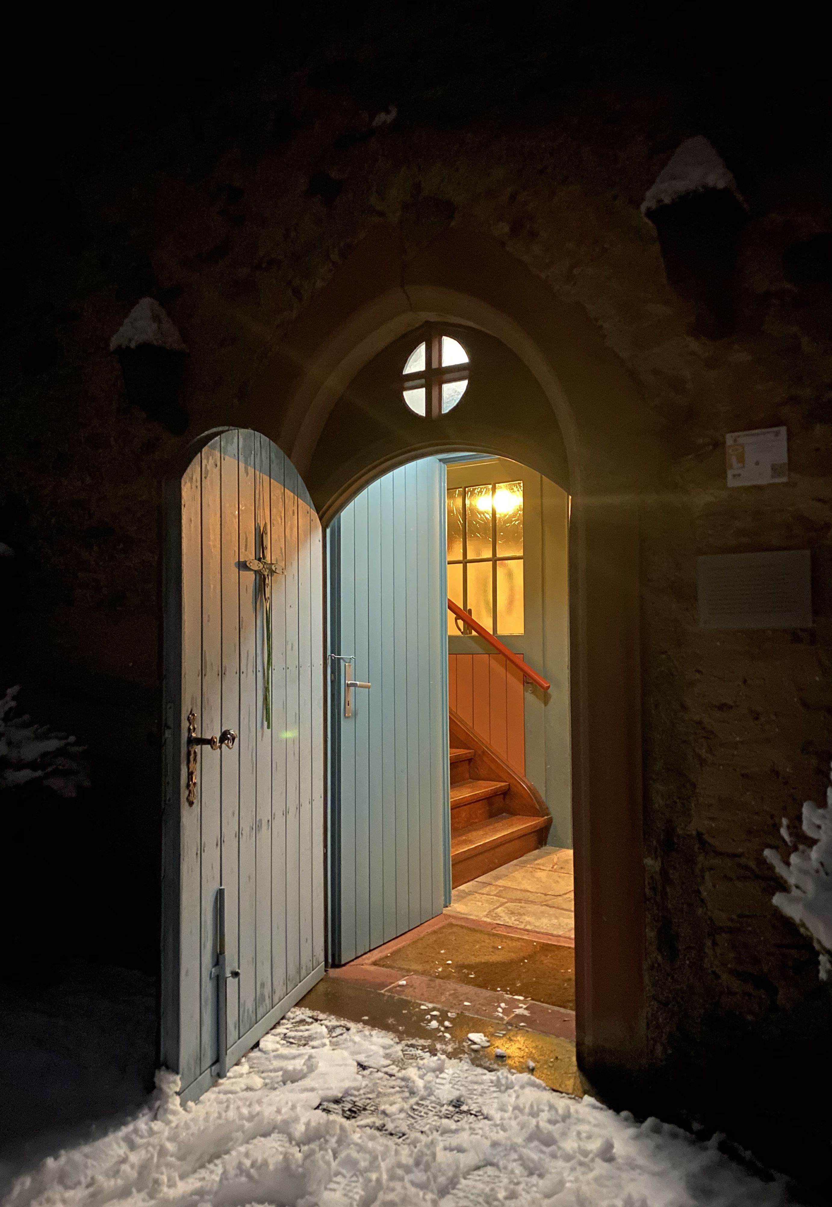 Eine geöffnete Holztür mit Kreuz führt von außen in einen beleuchteten Innenraum mit Treppe. Schnee liegt vor der Tür.