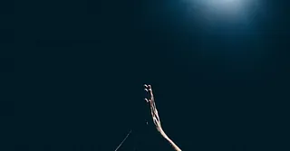 In der Dunkelheit recken sich zwei Hände empor. Sie sind zum Gebet aneinandergelegt. Von oben bescheint ein einzelnes Licht die Hände.