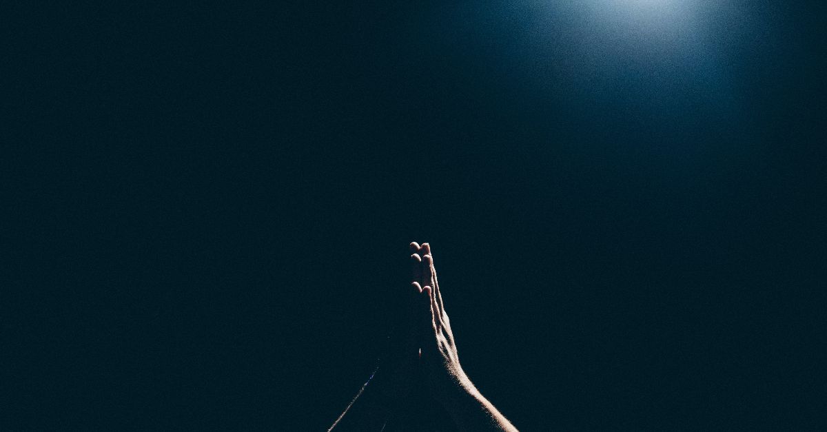 In der Dunkelheit recken sich zwei Hände empor. Sie sind zum Gebet aneinandergelegt. Von oben bescheint ein einzelnes Licht die Hände.