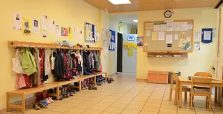 Flur des Kindergartens mit Garderobe, Jacken, Schuhen, Sitzbänken und einem Tisch mit Stühlen.