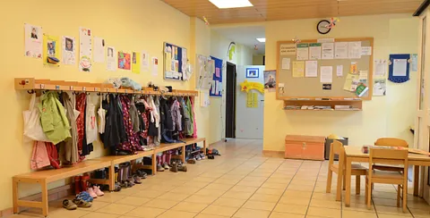 Flur des Kindergartens mit Garderobe, Jacken, Schuhen, Sitzbänken und einem Tisch mit Stühlen.