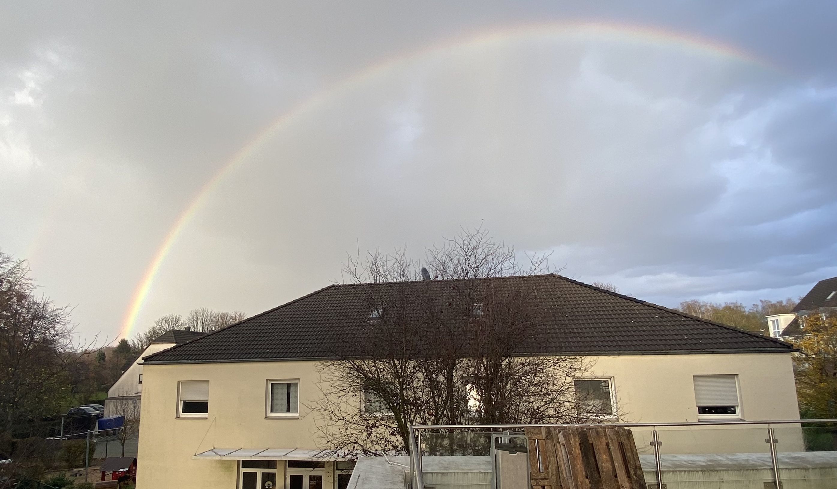 Regenbogen über dem zweistöckigen Kindergarten, mit einem kahlem Baum davor, unter bewölktem Himmel.