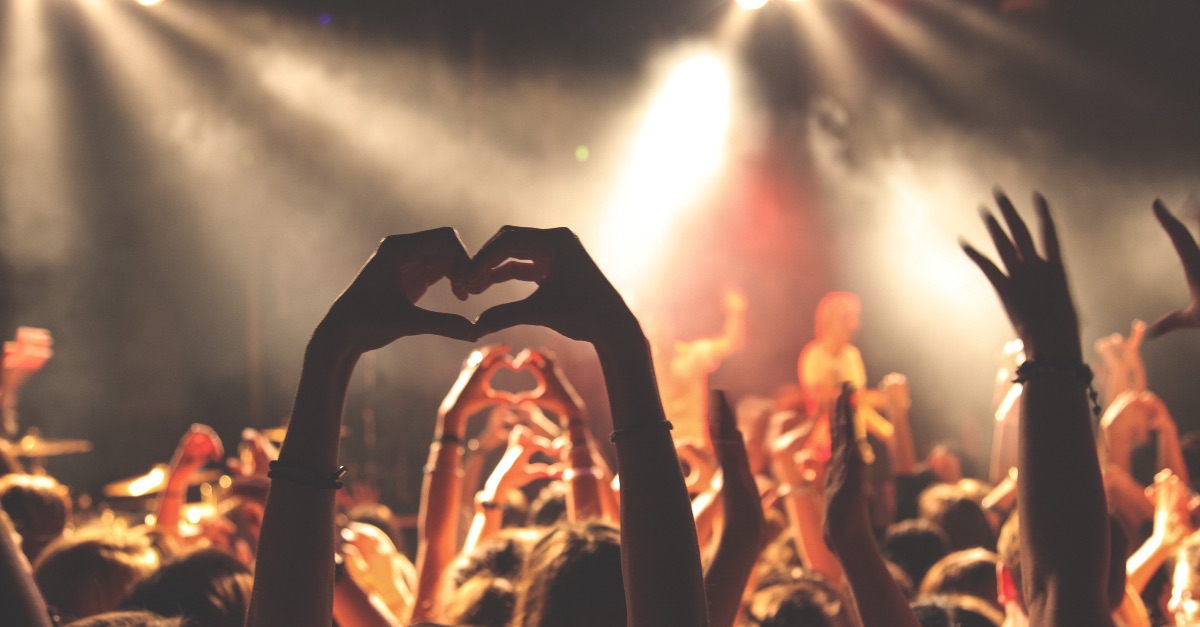Viele Menschen feiern bei einem Konzert, im Bildhintergrund zu sehen ist die Bühne. In der Menschenmenge hebt eine Person die Hände und formt ein Herz.