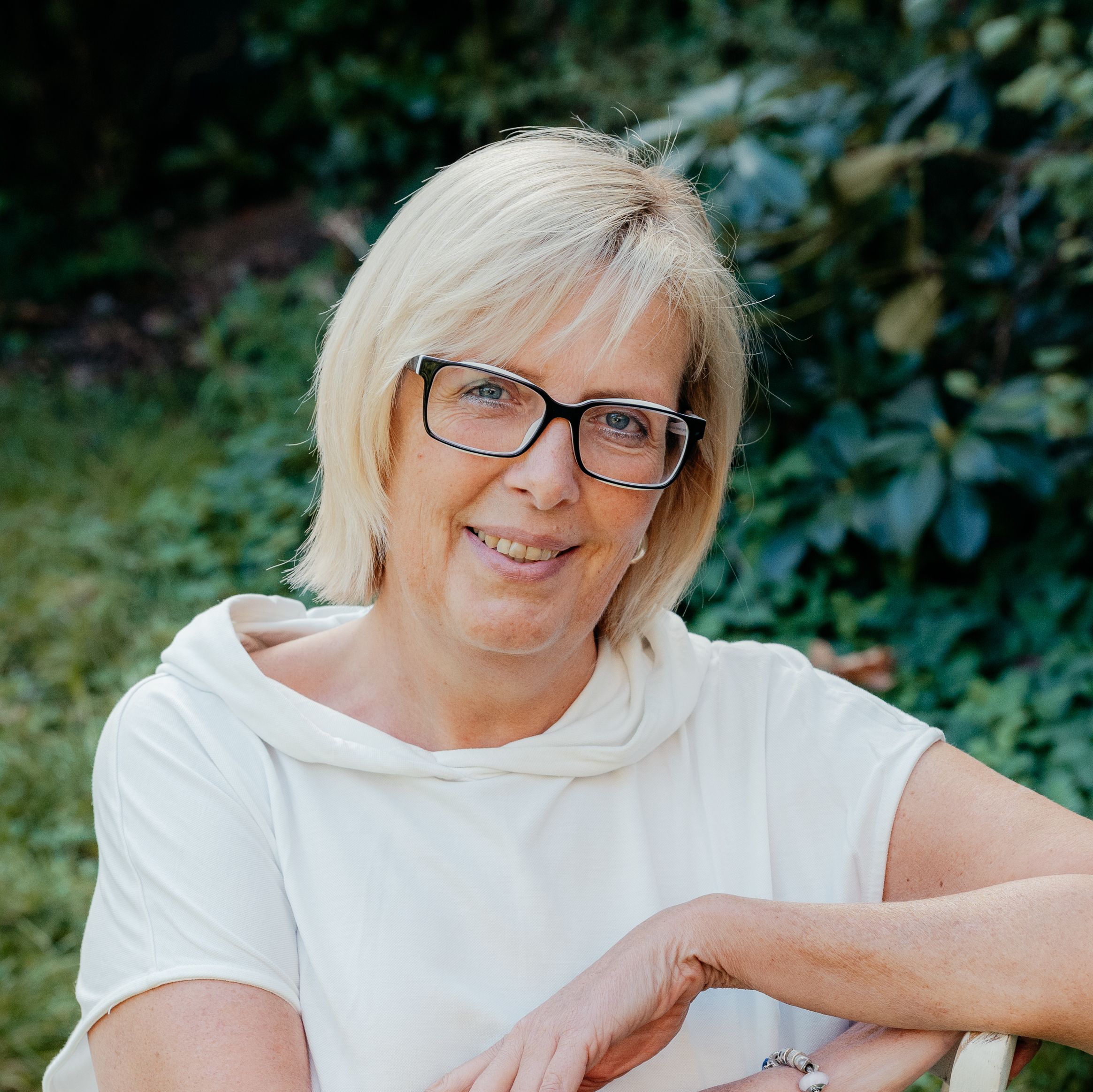 Portrait von Sonja Zickwolf-Krebs, sitzt entspannt auf einem weißen Stuhl im Garten, lächelt.