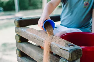Ein Kind gießt Sand aus einem blauen Becher auf eine hölzerne Sandspielstruktur im Freien.