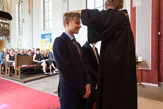 Konfirmand im Anzug erhält im Gottesdienst im Kirchenraum den Segen von einer Pfarrerin im Talar. Im Hintergrund sitzt die Gemeinde in den Bänken.