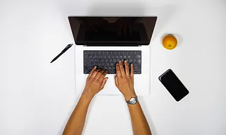 Hände tippen auf einer Laptop-Tastatur, daneben liegen Stift, Orange und Smartphone auf weißem Tisch.