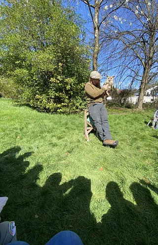 Auf einer Wiese steht ein Mann. Auf seiner Hand sitzt ein Wildvogel. Im Bildvordergrund zu sehen sind die Schatten von Kindern, die ihm zusehen.