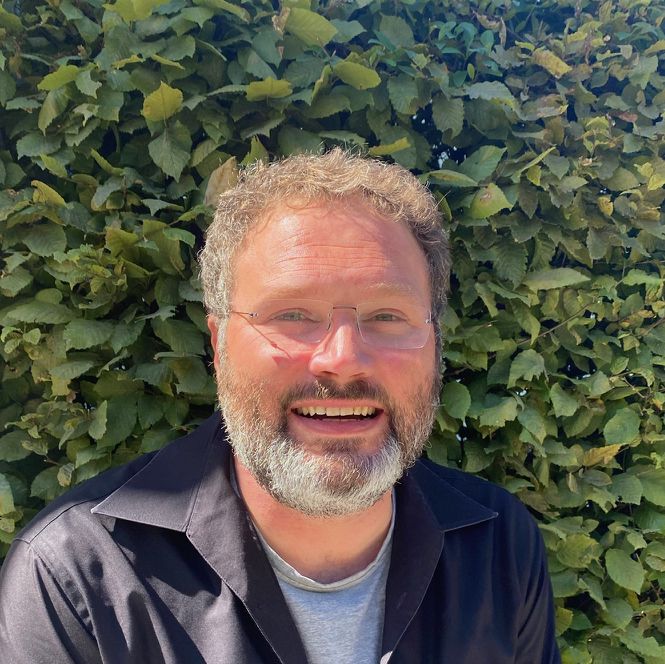 Portrait von Michael Lavista, sitzt lächelnd vor einem grünen Busch, trägt schwarze Hemd und graues T-Shirt.