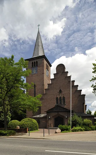Adolf-Clarenbach-Kirche mit steilem Turm und Treppengiebel aus rotem Backstein, umgeben von Bäumen.