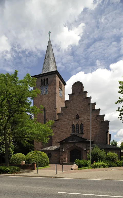 Adolf-Clarenbach-Kirche mit steilem Turm und Treppengiebel aus rotem Backstein, umgeben von Bäumen.