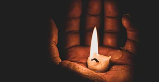In der Dunkelheit ist eine Hand zu sehen. Die nach oben geöffnete Hand hält eine heruntergebrannte Kerze, die die Hand beleuchtet.