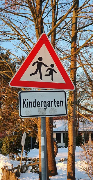 Ein Verkehrsschild mit rotem Dreieck und Symbol von zwei Kindern. Darunter ein Schild mit der Aufschrift "Kindergarten", im winterlichen Umfeld mit Bäumen und Schnee.