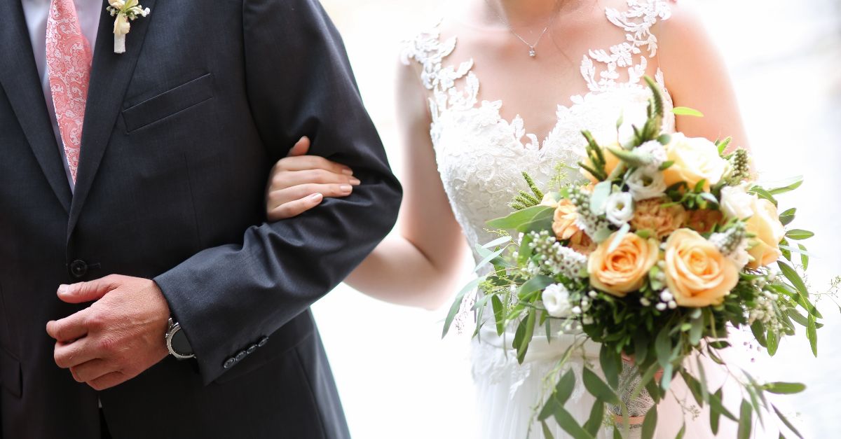Eine Braut in weißem Spitzenkleid hält einen Blumenstrauß mit gelben Rosen und grünen Blättern, Arm in Arm mit dem Bräutigam im schwarzen Anzug und rosa Krawatte.