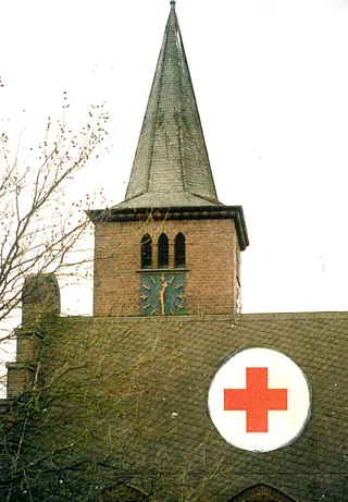 Kirchturm der Adolf-Clarenbach-Kirche, mit spitzem Dach, davor das Dach des Kirchenschiffs mit dem Schutzzeichen rotes Kreuz auf weißem Grund.