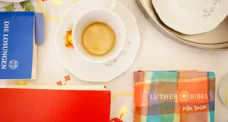 Tisch mit einer Tasse Kaffee, einer bunten Lutherbibel, einem roten (Gesang-)Buch und einem Büchlein mit den Herrnhuter Losungen.