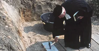 In einer Baugrube haut ein Pfarrer im Talar und mit Barett mit einem Hammer auf den Grundstein vor Baubeginn.