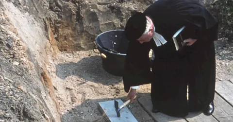 In einer Baugrube haut ein Pfarrer im Talar und mit Barett mit einem Hammer auf den Grundstein vor Baubeginn.