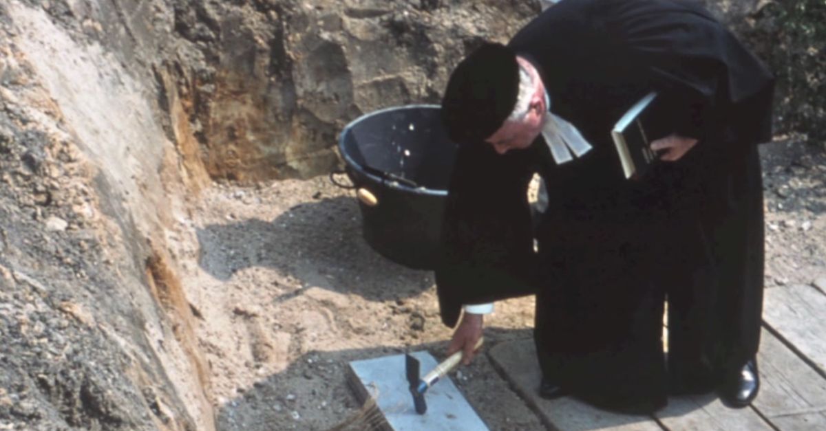 In einer Baugrube haut ein Pfarrer im Talar und mit Barett mit einem Hammer auf den Grundstein vor Baubeginn.