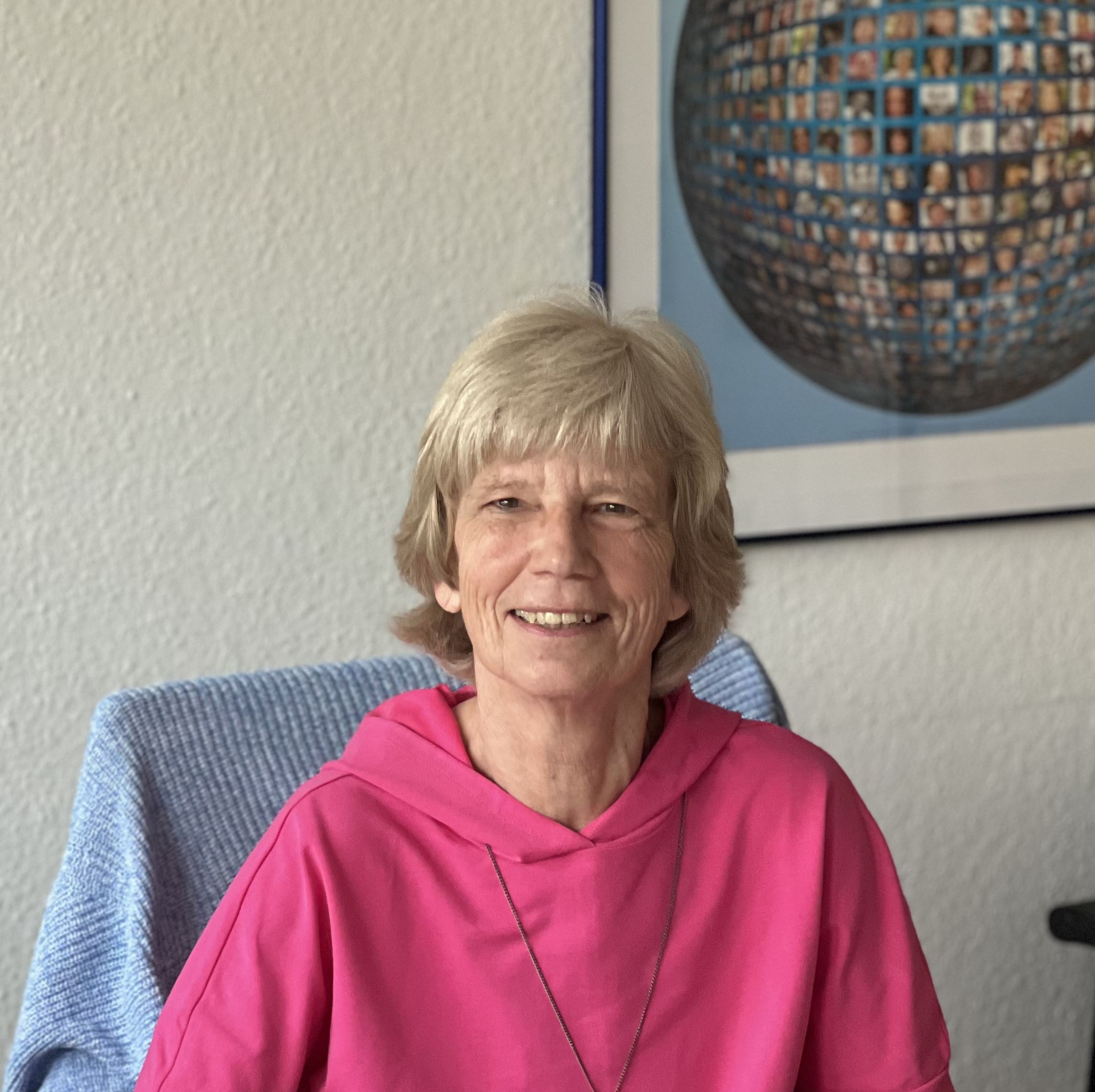 Portrait von Petra Röder, lächelnd, am Schreibtisch mit pinkem Ordner, im Hintergrund ein Kunstbild an der Wand.