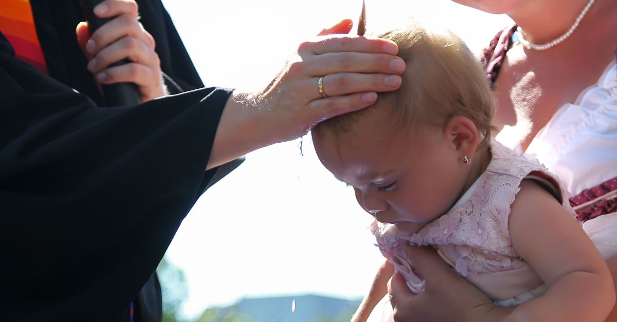 Ein Baby wird bei einer Taufe vom Pfarrer gesegnet, während es von einer anderen Person gehalten wird. Wasser tropft herab.