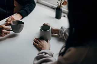 Zwei Menschen sitzen im Gespräch an einem Tisch. Sie halten jeweils eine Tee- oder Kaffeetasse in der Hand.