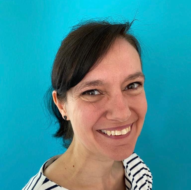 Portrait von Dana Rupieper, lächelnd, trägt ein weißes Shirt mit schwarzen Streifen vor blauem Hintergrund.